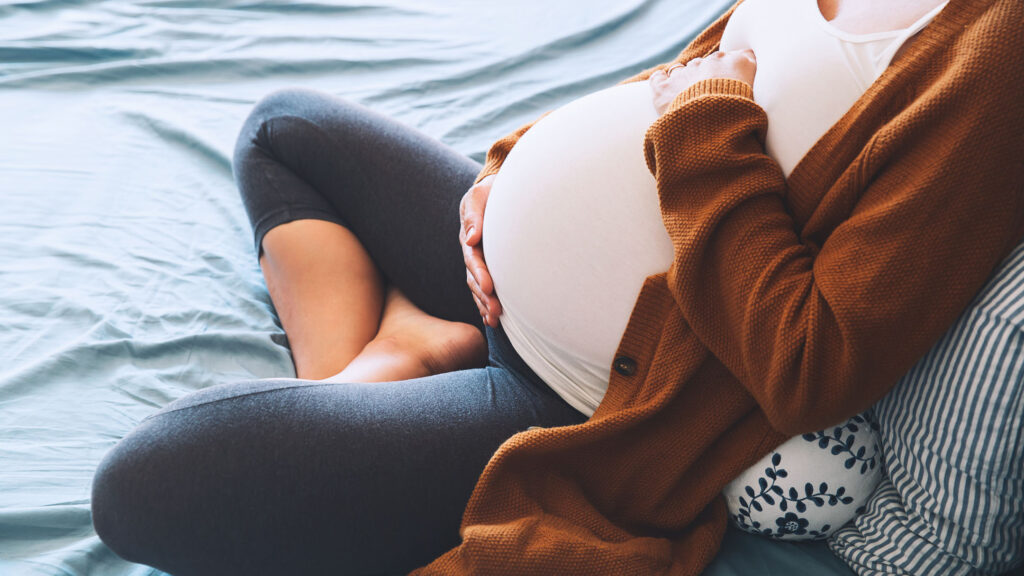 pregnant woman sitting at bed and holds hands on belly in bedroom at home.