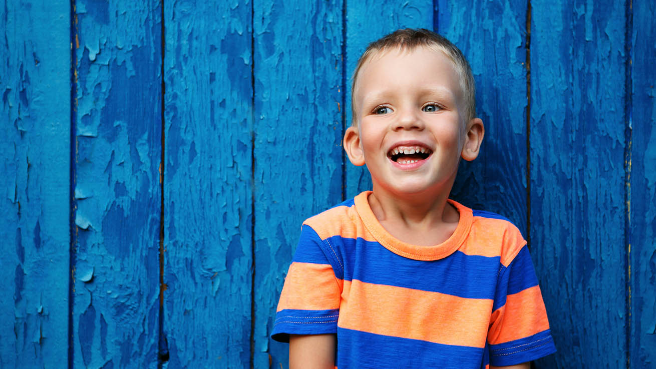 Smiling boy standing in front of a fence that has peeling paint.