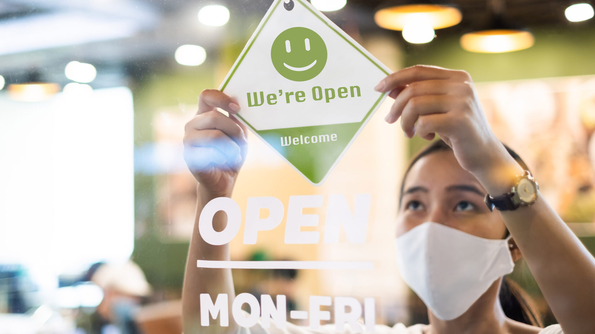 A woman wearing a mask hangs an open sign on the door of a cafe