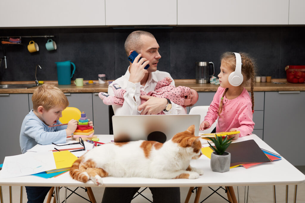 Chaotic scene of a dad working from home while holding a baby and caring for two young children