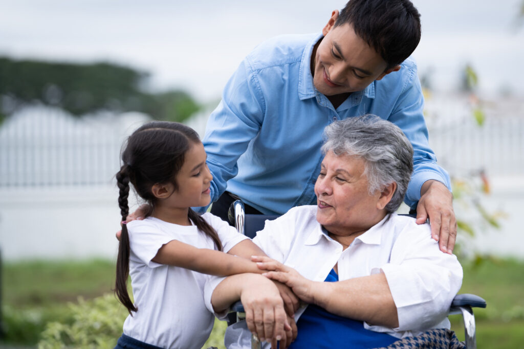 A family consisting of a man, young girl, and older woman in a wheelchair smiling and talking 