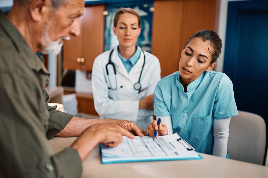 a nurse assisting a senior man to fill out medical documents at reception desk at doctor's office.