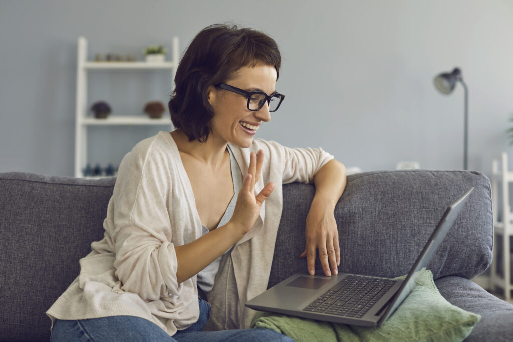 smiling woman sitting at home with laptop and greeting somebody during online conversation or videocall. 