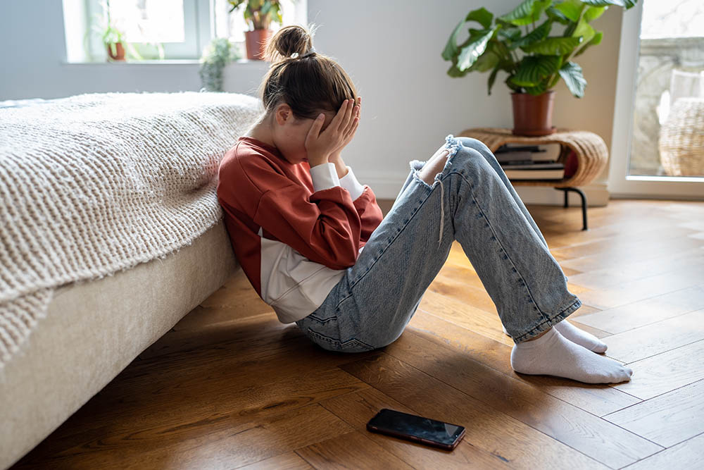 Teen girl covering face with hands and crying while sitting on floor with mobile phone nearby