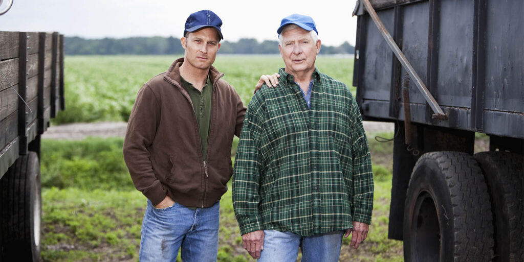 A senior man and his adult son working on the family farm. In the background is a field of crops.