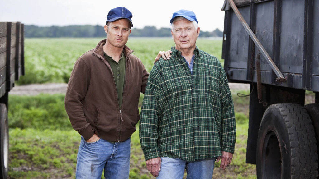 A senior man and his adult son working on the family farm, growing potatoes. They are standing between two trucks, with serious expressions, looking at the camera. The younger man is resting his hand on his father's shoulder, and hand the other in the pocket of his jeans. He is wearing a brown jacket. The older man is wearing a green, plaid shirt. In the background is a field of crops.