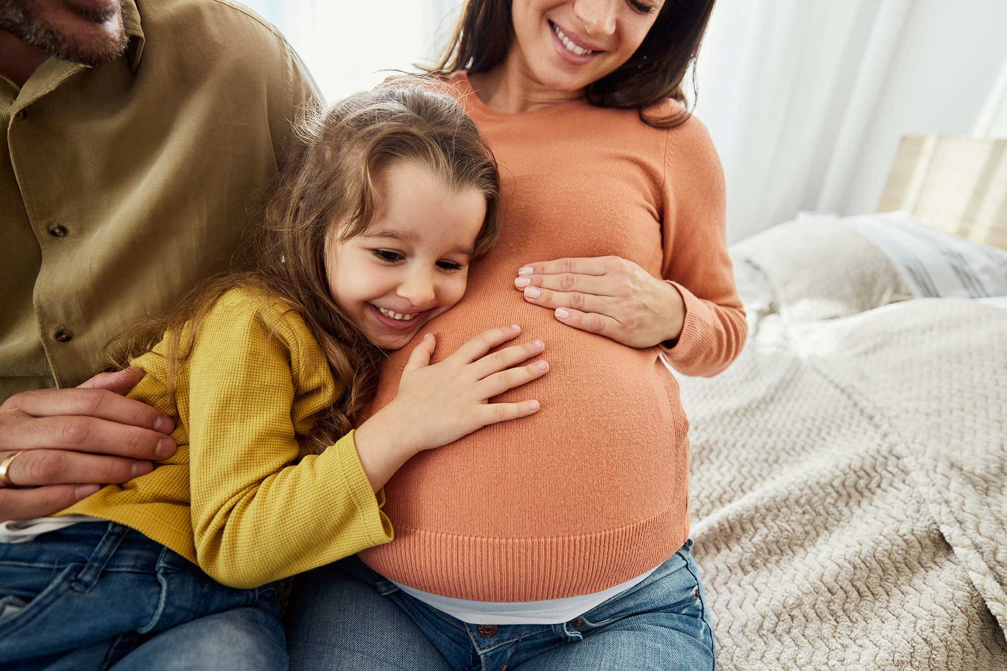 A happy father, mother, and daughter sit on a bed and embrace, with the daughter holding the mother's pregnant belly.